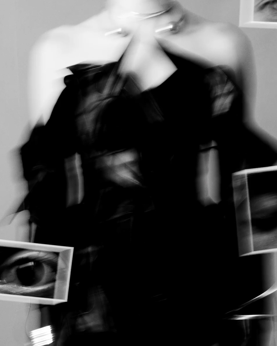 Blurred image of a person in a black dress and silver necklace, with framed eye images as props in a muted indoor setting.