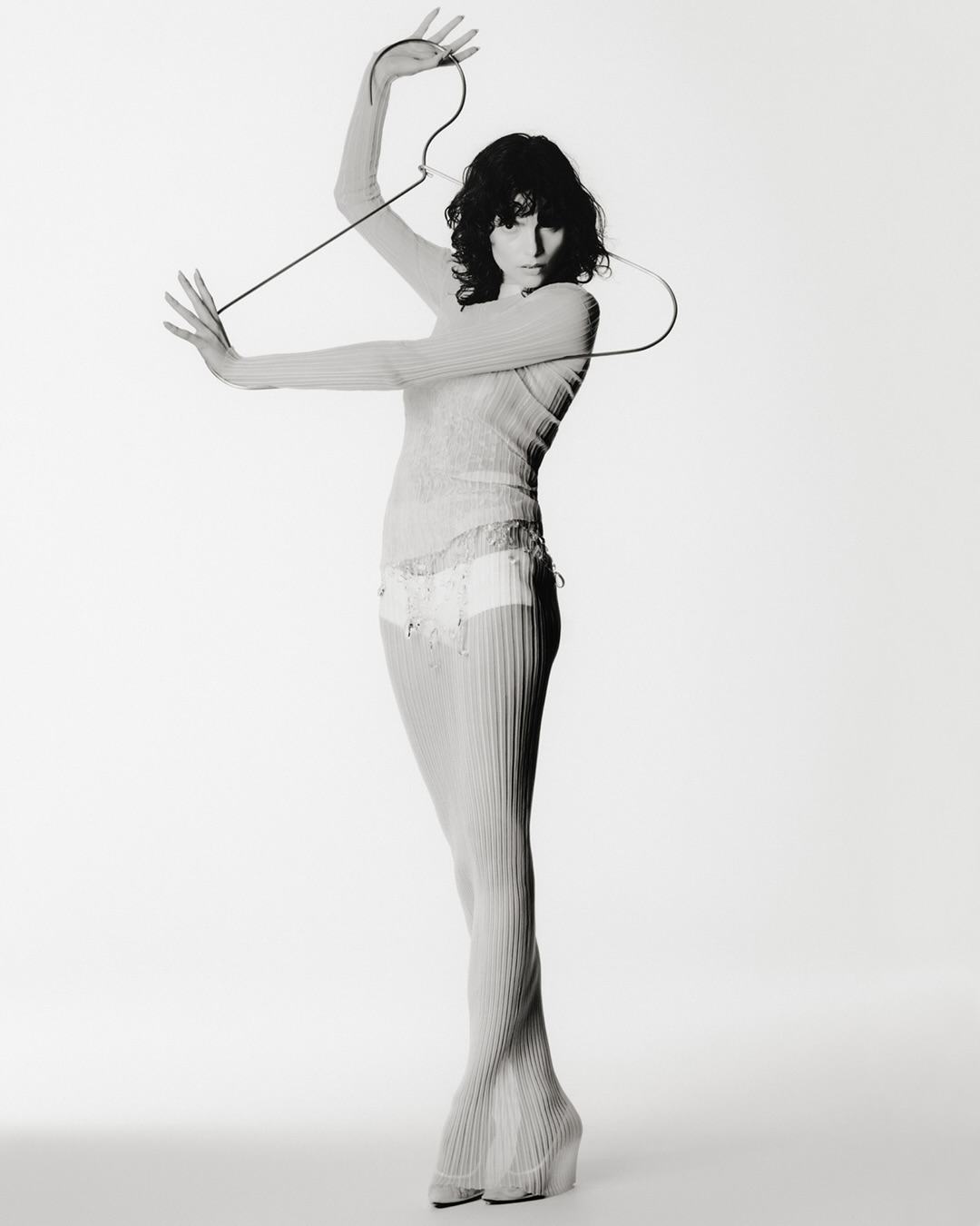 Monochrome full shot of a woman in sheer knitwear holding a wire hanger against a white background in a fashion editorial setting.