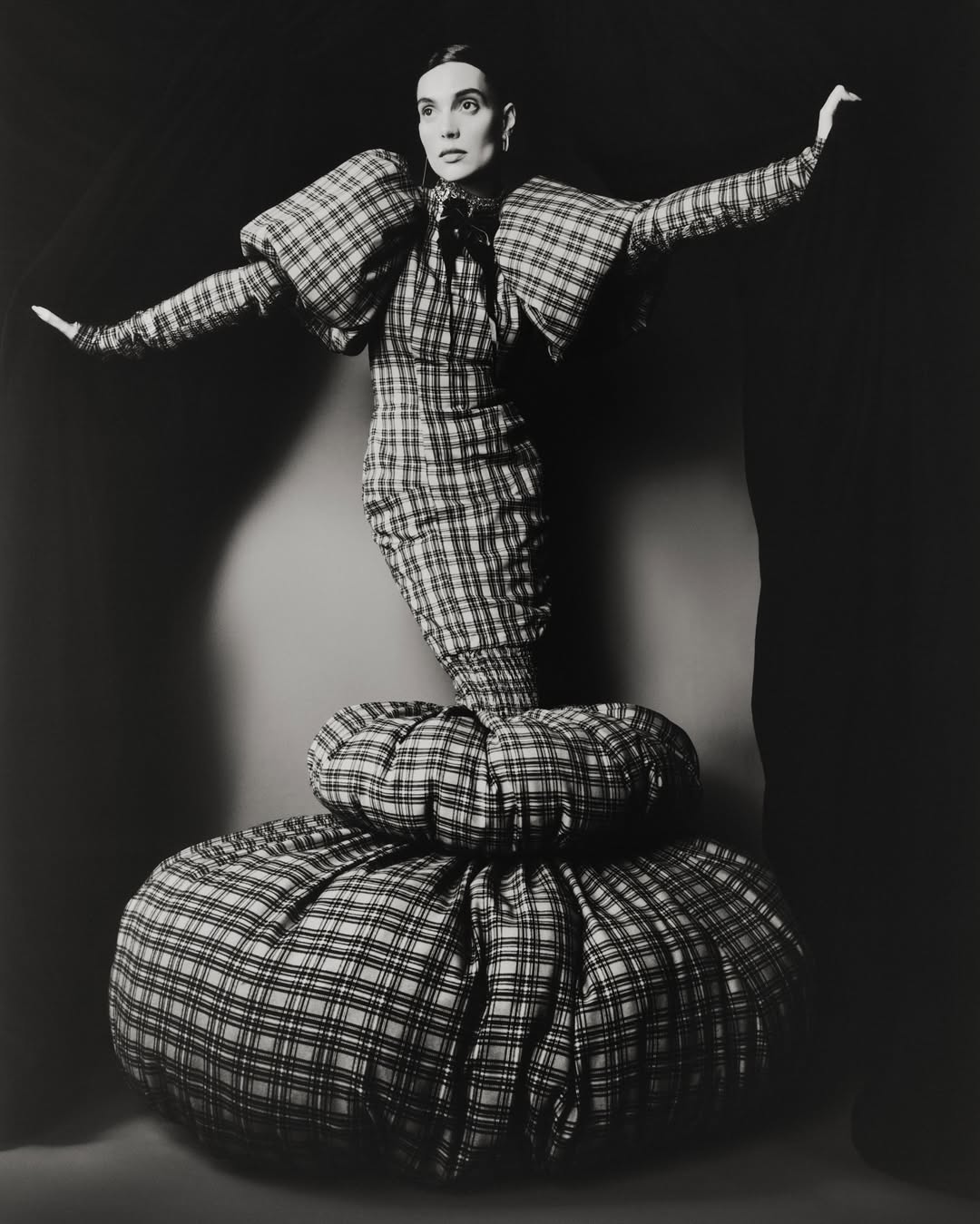 Monochrome studio shot of a woman in a plaid dress with voluminous shoulders and skirt, arms outstretched against a dark background.