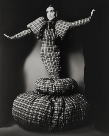 Monochrome studio shot of a woman in a plaid dress with voluminous shoulders and skirt, arms outstretched against a dark background.