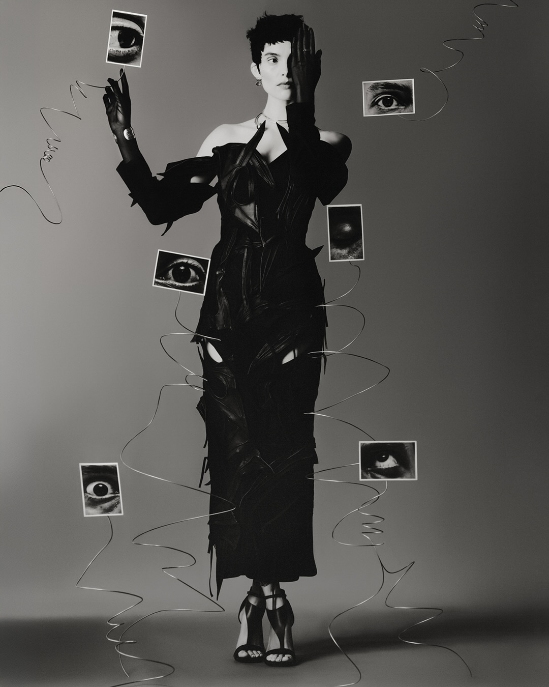 Monochrome fashion shot of a woman in a dark, avant-garde dress surrounded by eye photographs, conveying a surreal, artistic aesthetic.