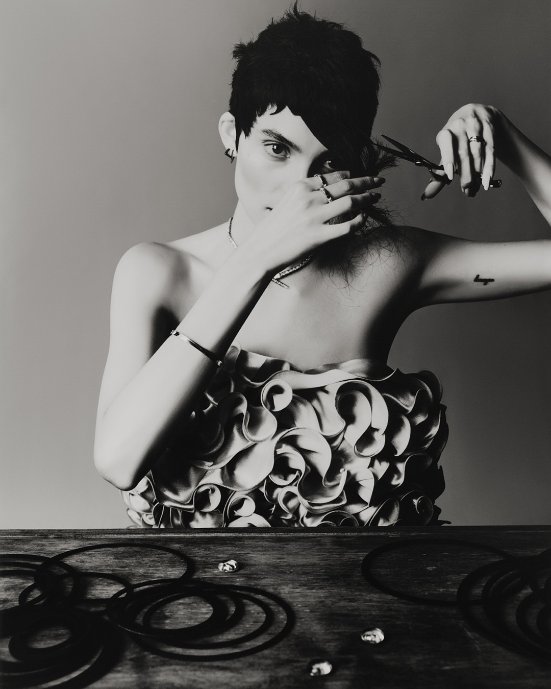 Monochrome portrait of a shirtless person with short hair, cutting their own hair with scissors above a wood table with hair ties.