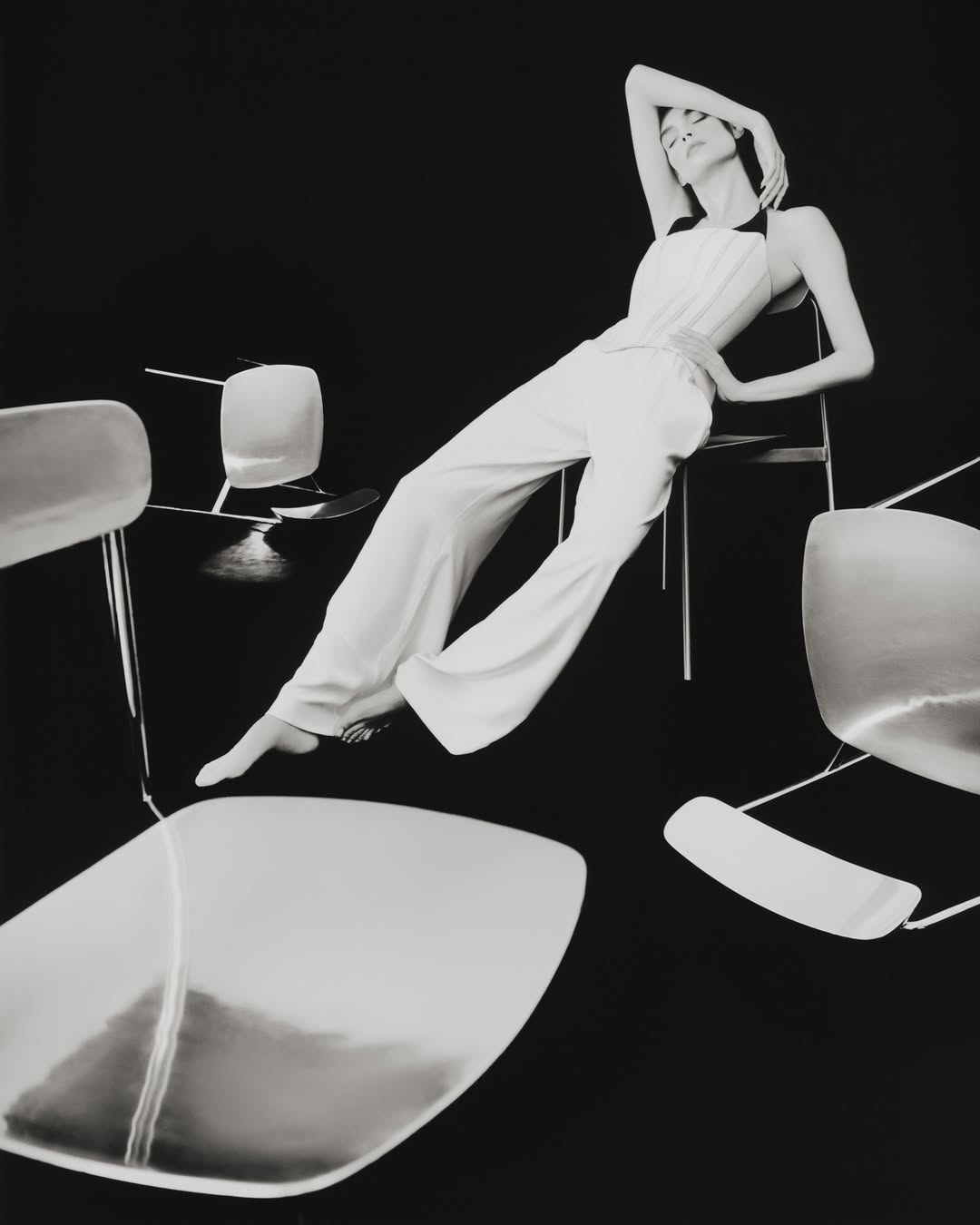Monochromatic fashion shot of a woman in a white halter jumpsuit lounging on a chair, surrounded by floating chairs against a black background.