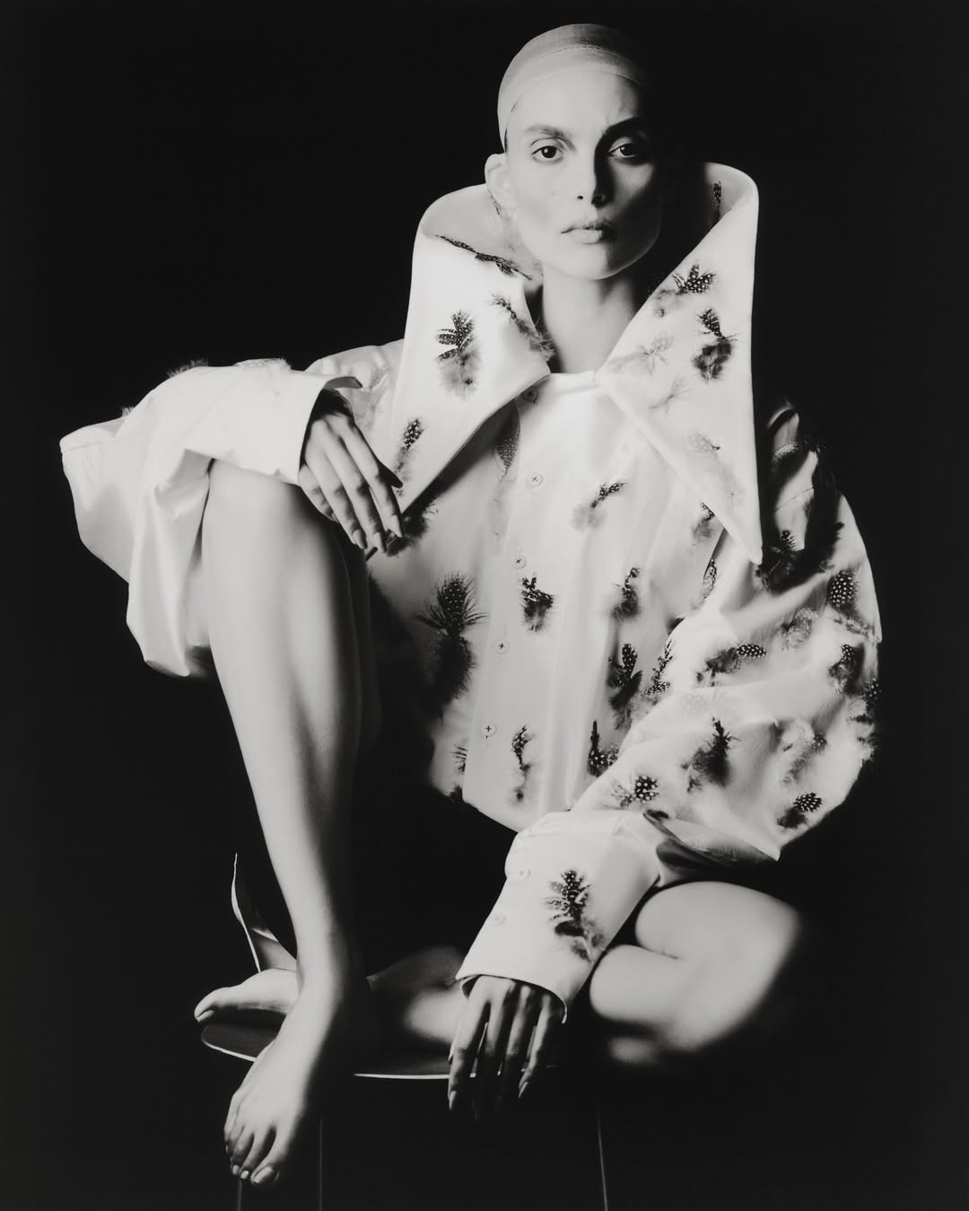 Striking black and white fashion portrait of a seated person with a shaved head, wearing an oversized collared shirt with a botanical print.