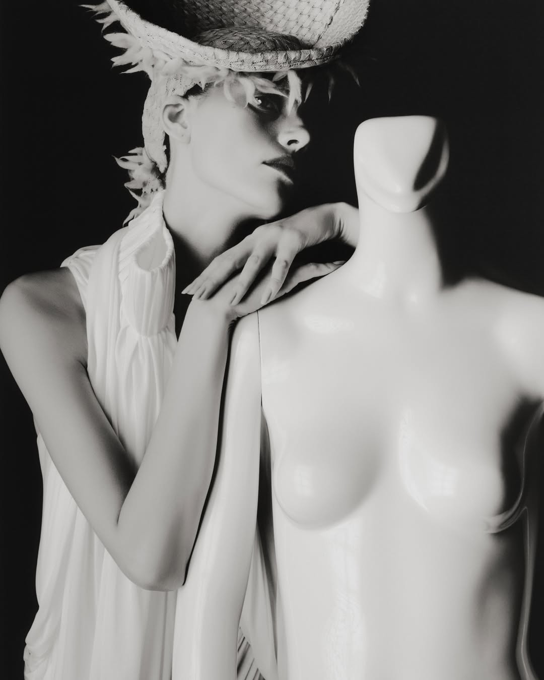 A model with a decorative hat poses with a mannequin in a studio, black and white fashion portrait. The lighting is bright, and the background is dark.