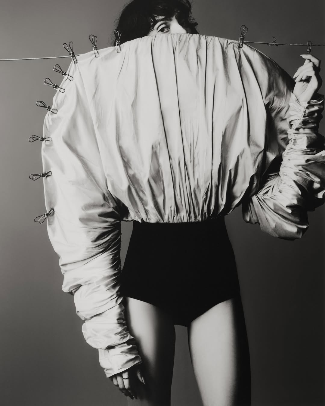 A woman poses in a studio wearing a voluminous, draped jacket secured with clothespins, paired with a high-waisted black garment.