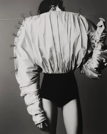 A woman poses in a studio wearing a voluminous, draped jacket secured with clothespins, paired with a high-waisted black garment.
