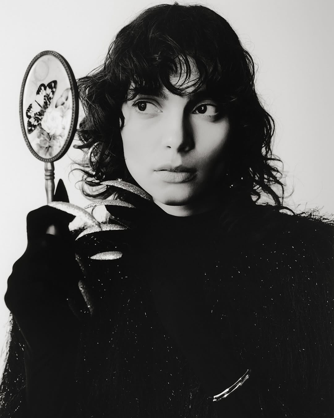 A portrait of a brunette woman holding a vintage mirror with a butterfly design, against a plain white backdrop, close-up.