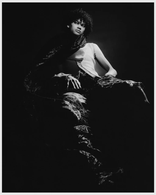Monochrome fashion shot of an adult female posing with a scarf, wearing a top and patterned bottoms, set against a dark background.