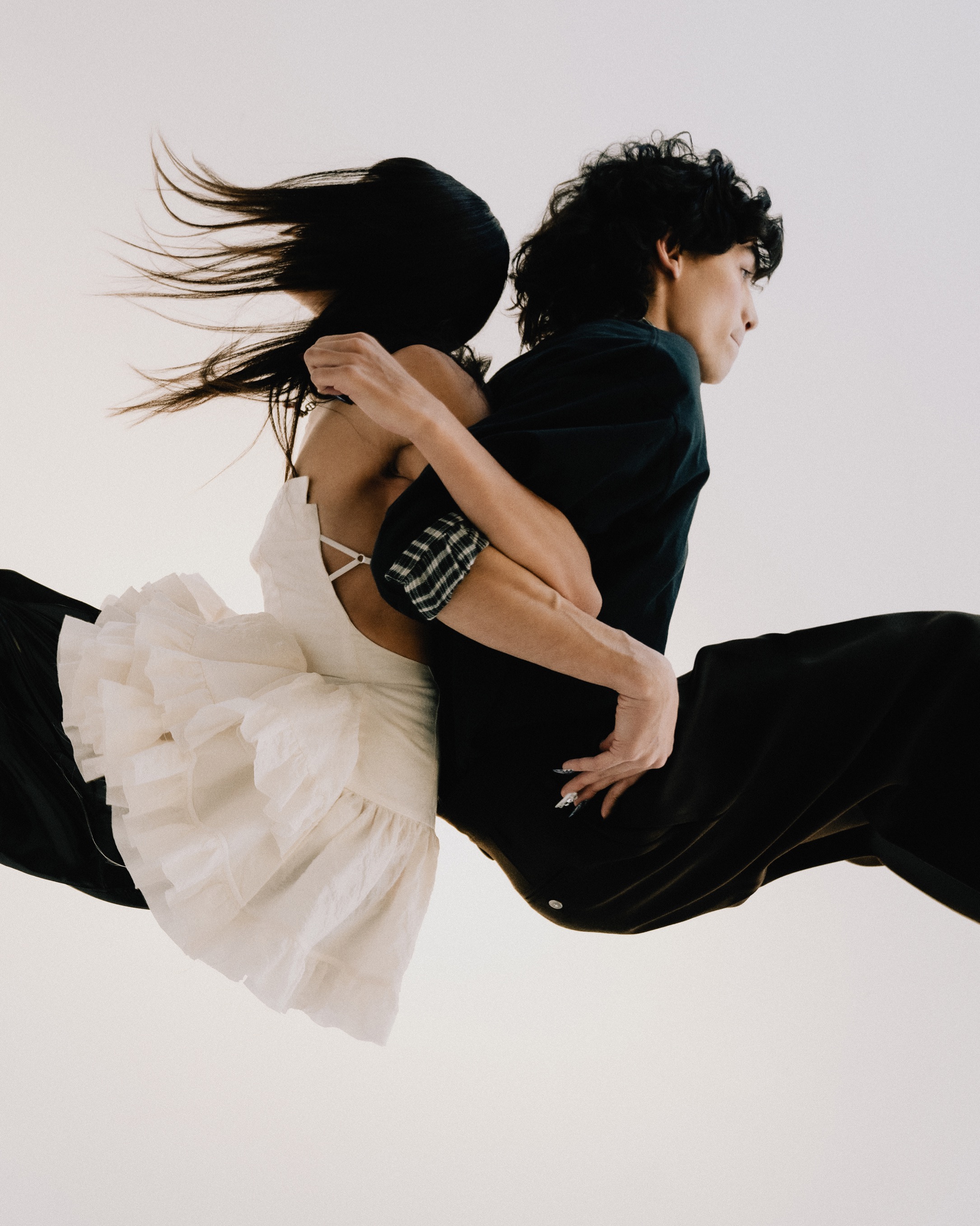 A couple embraces in mid-air, the woman in a ruffled white dress and the man in black, set against a bright, neutral background.
