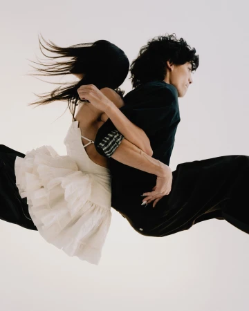 A couple embraces in mid-air, the woman in a ruffled white dress and the man in black, set against a bright, neutral background.