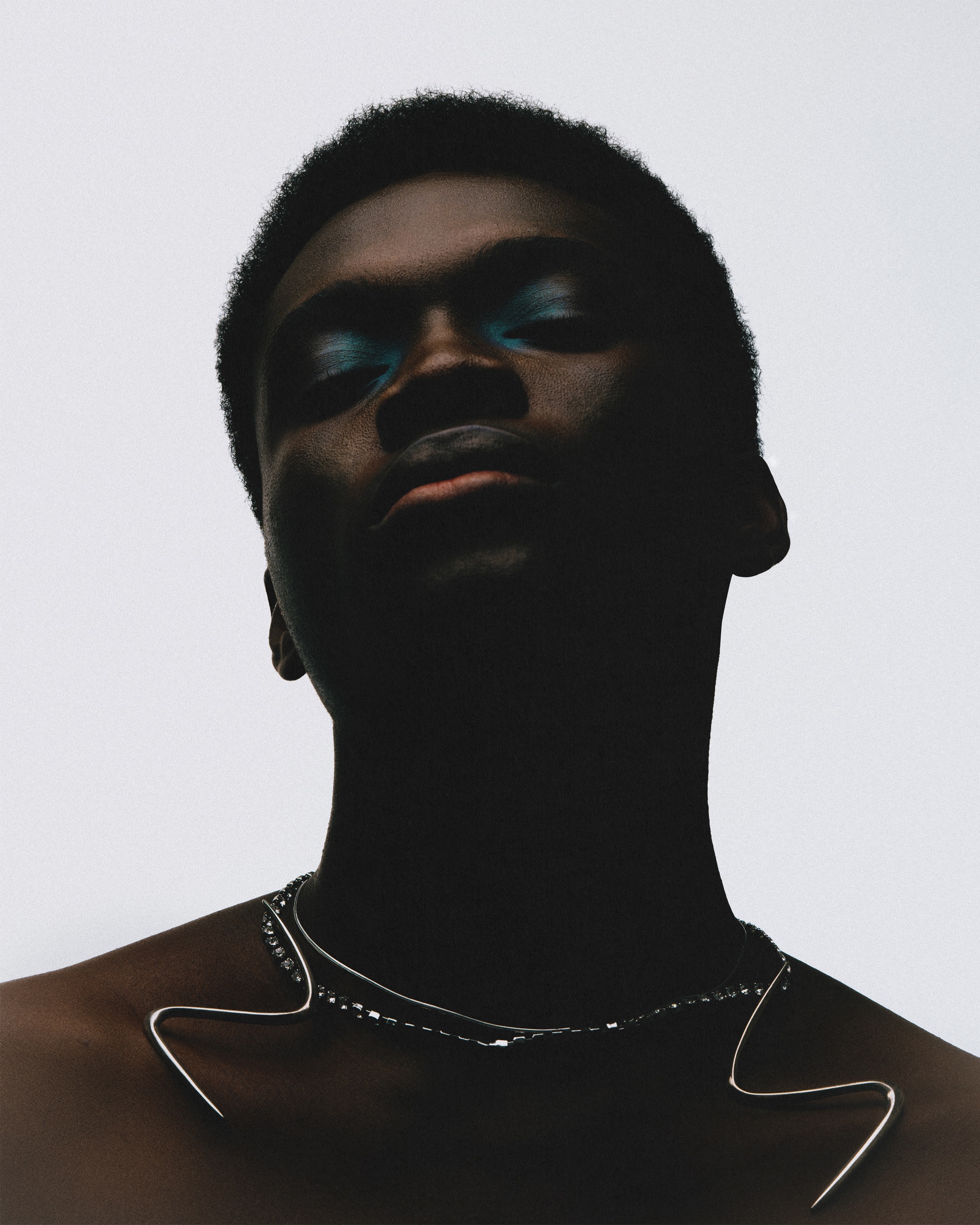 Headshot of a young adult with dark skin and teal eyeshadow, wearing a silver necklace, against a stark white background.