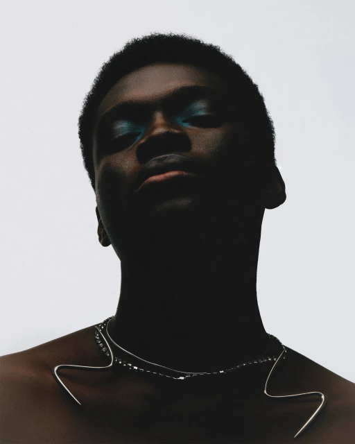 Headshot of a young adult with dark skin and teal eyeshadow, wearing a silver necklace, against a stark white background.
