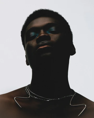 Headshot of a young adult with dark skin and teal eyeshadow, wearing a silver necklace, against a stark white background.