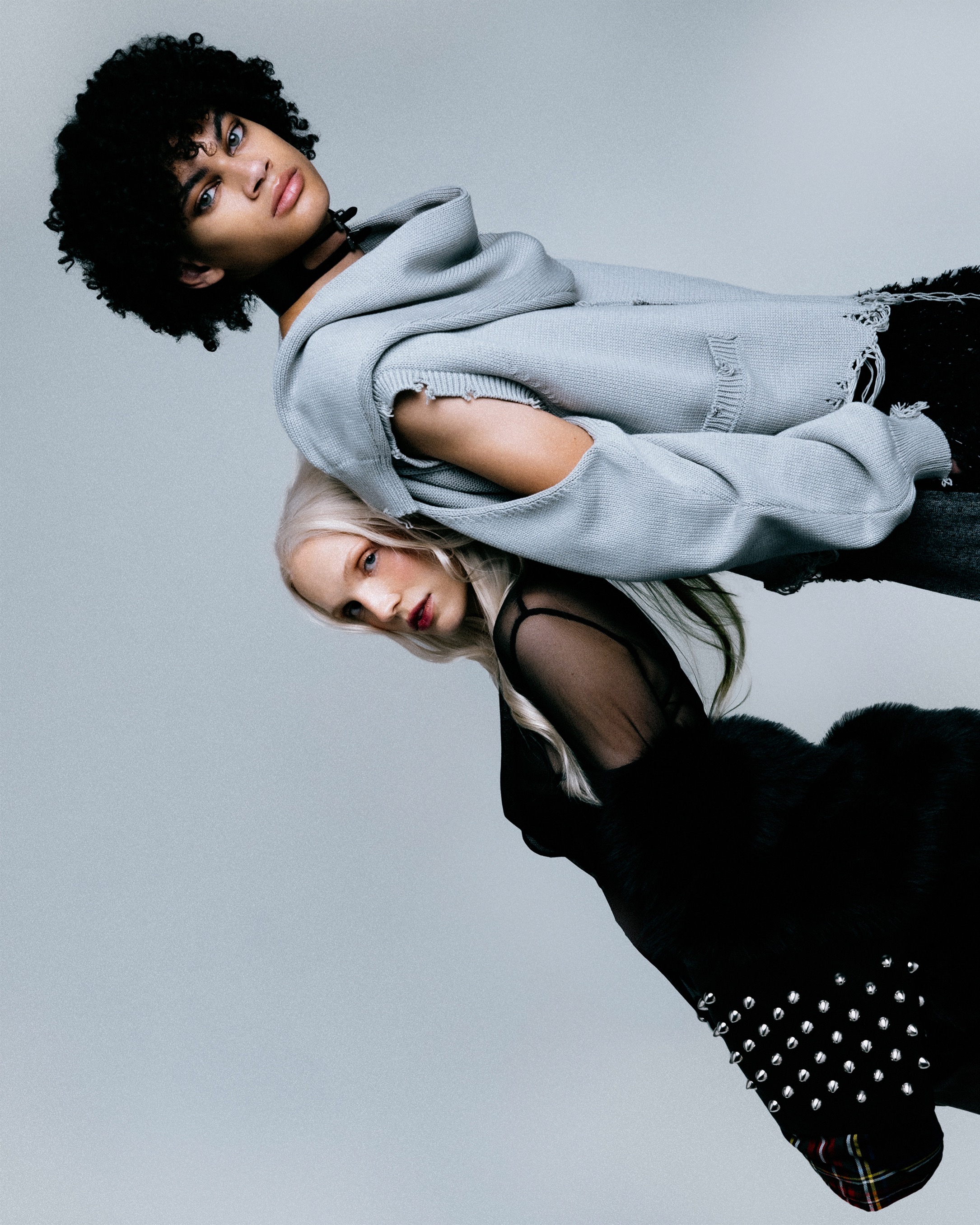 Two fashion models pose in a studio portrait. One with dark, curly hair wears a light blue hoodie; the other has blonde hair and a black dress.