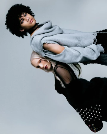 Two fashion models pose in a studio portrait. One with dark, curly hair wears a light blue hoodie; the other has blonde hair and a black dress.