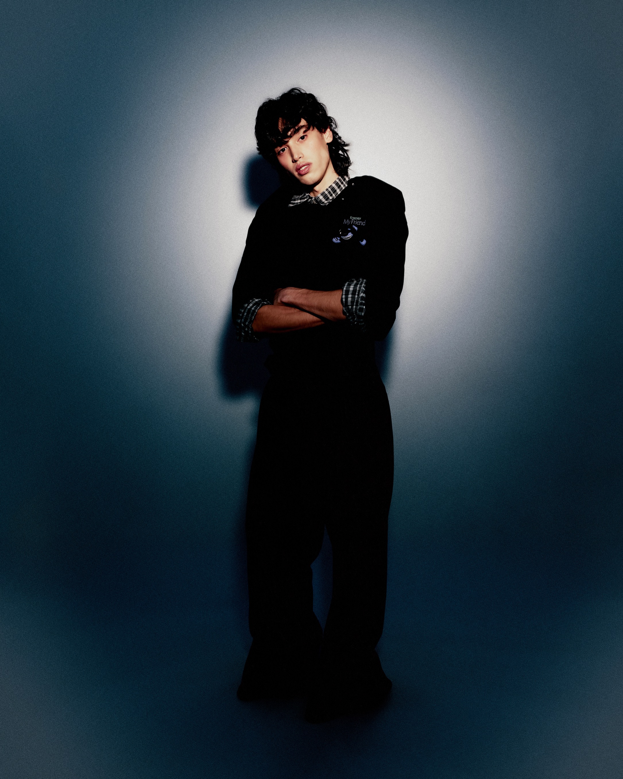 A young man with long dark hair poses in a black suit against a gradient grey backdrop in a full studio shot, arms crossed.
