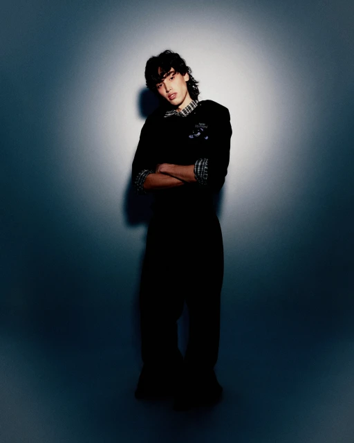 A young man with long dark hair poses in a black suit against a gradient grey backdrop in a full studio shot, arms crossed.