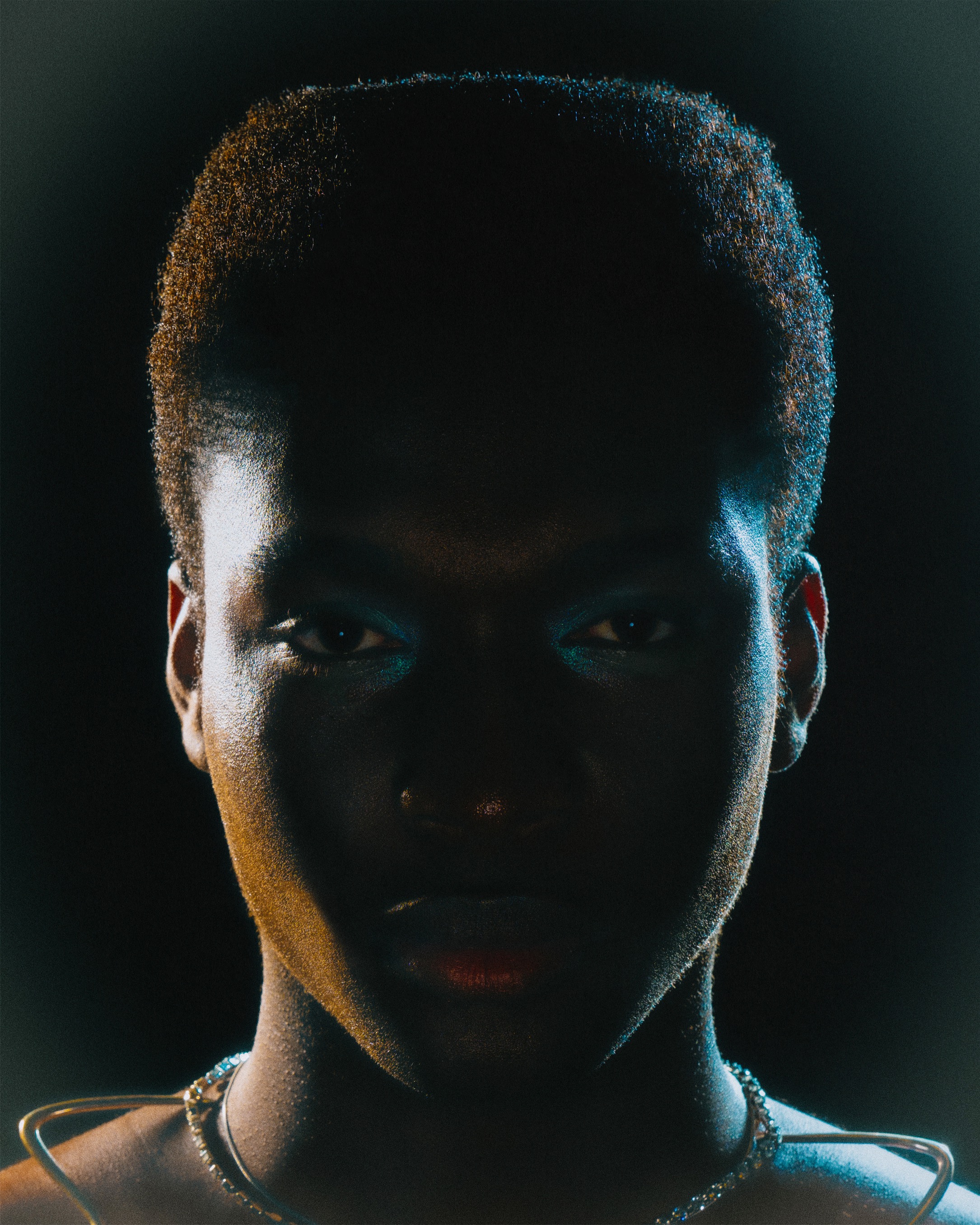 Close-up portrait of an adult, dark-skinned woman with an intense gaze, illuminated by vibrant teal and yellow lighting against a dark background.