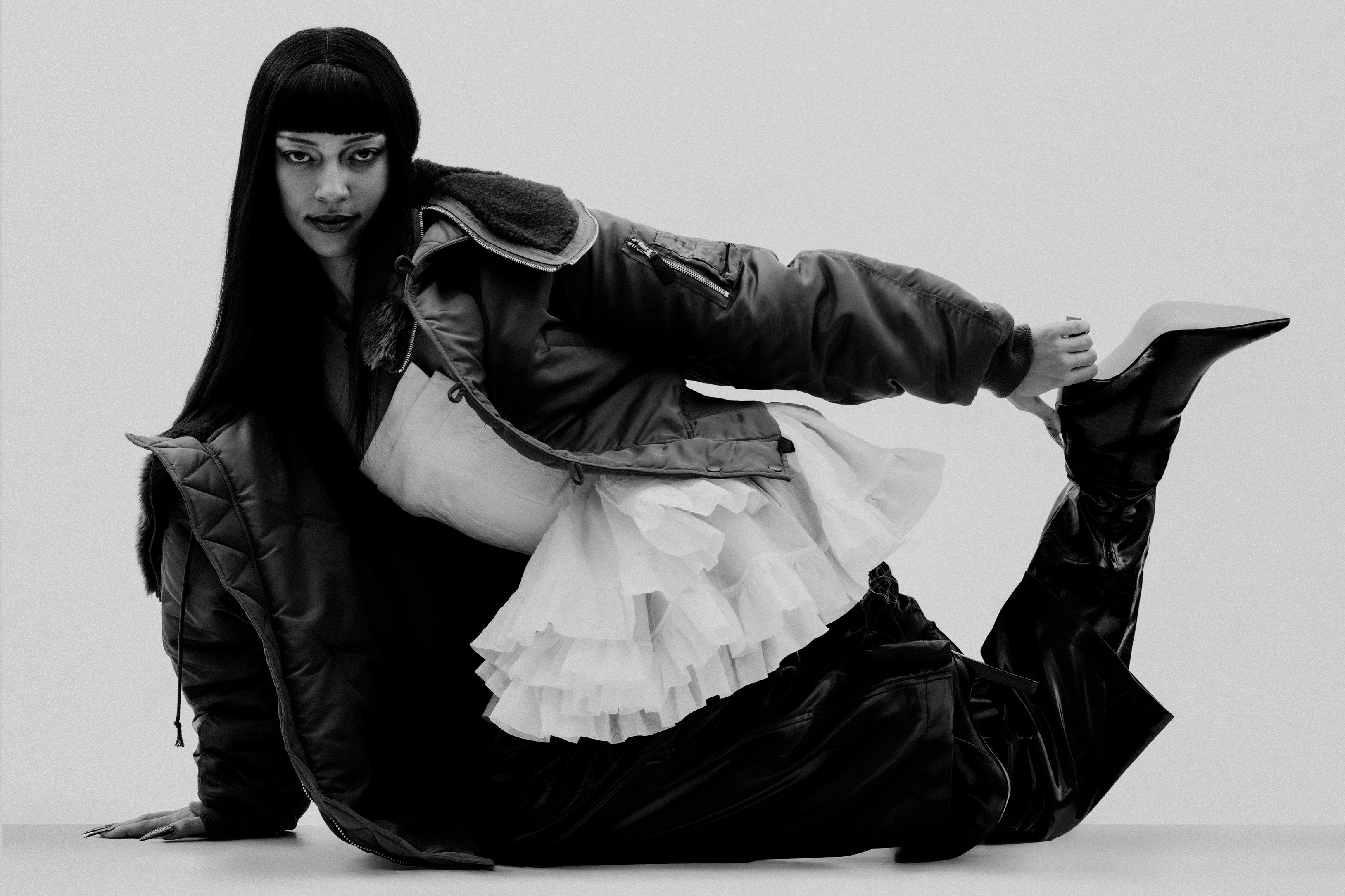 Woman poses in black jacket and black patent boots against white background. High fashion portrait. Moody lighting. Fashion editorial.