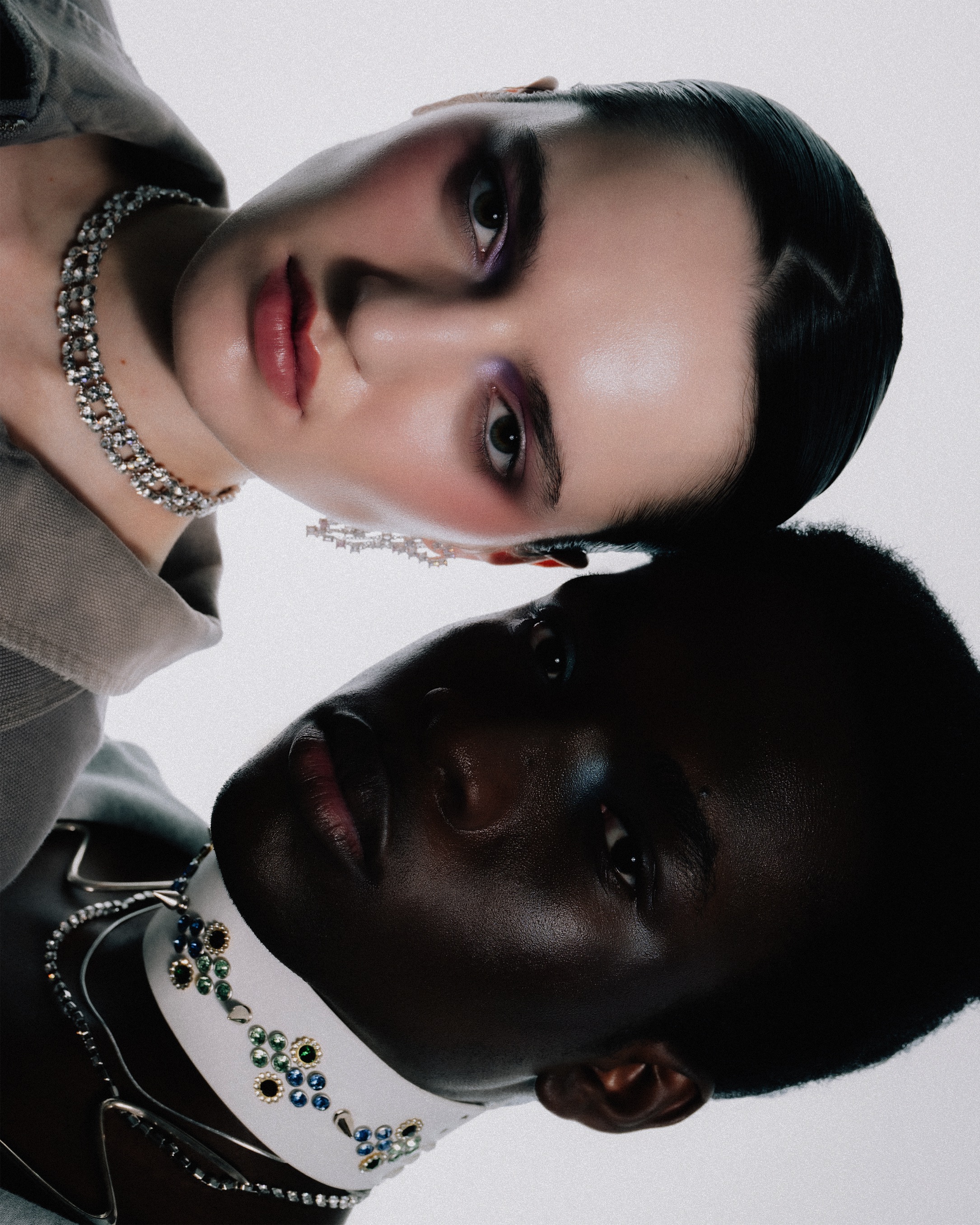Close-up of two fashion models, one fair-skinned and one dark-skinned, wearing jewelry and makeup against a bright white background.