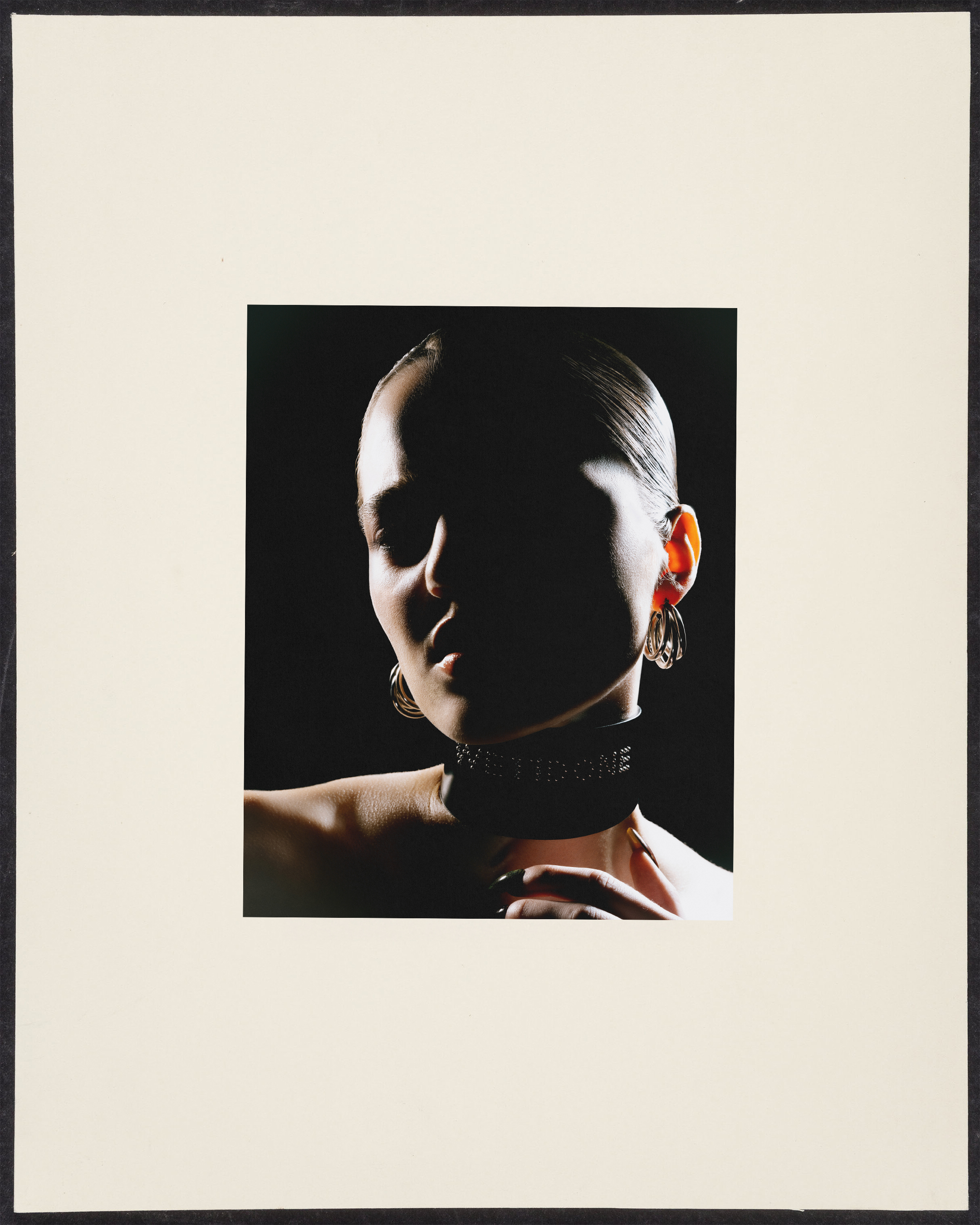 Close-up portrait of a woman with slicked-back hair wearing a black choker and teardrop earrings against a stark black background.