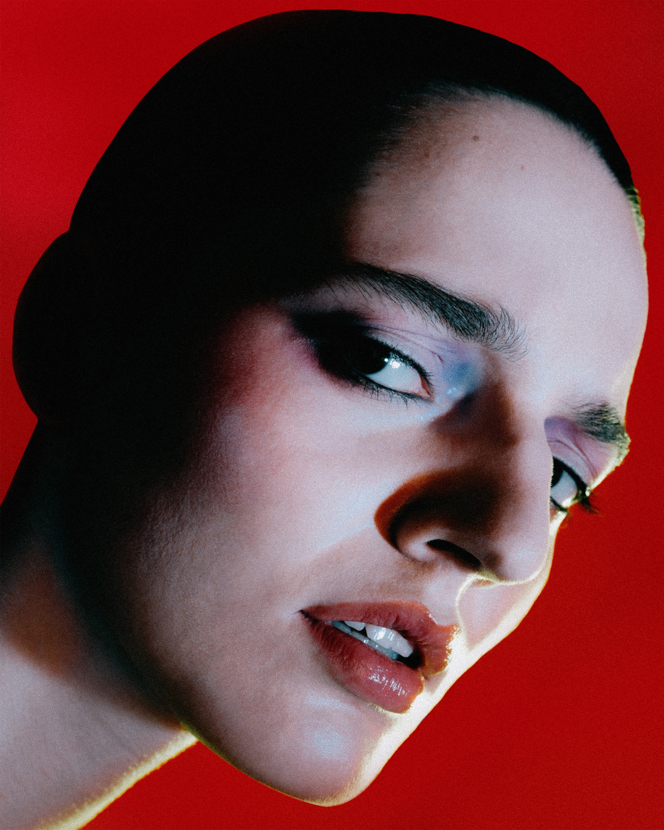 Close-up portrait of a woman with glossy lips and blue eyeshadow against a bright red background, angled for beauty or fashion content.