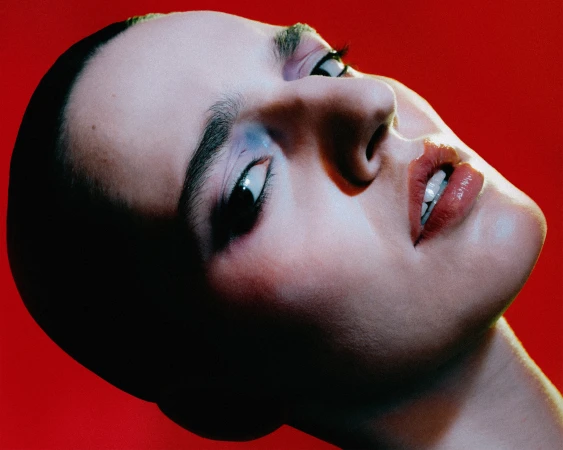 Close-up portrait of a woman with glossy lips and blue eyeshadow against a bright red background, angled for beauty or fashion content.