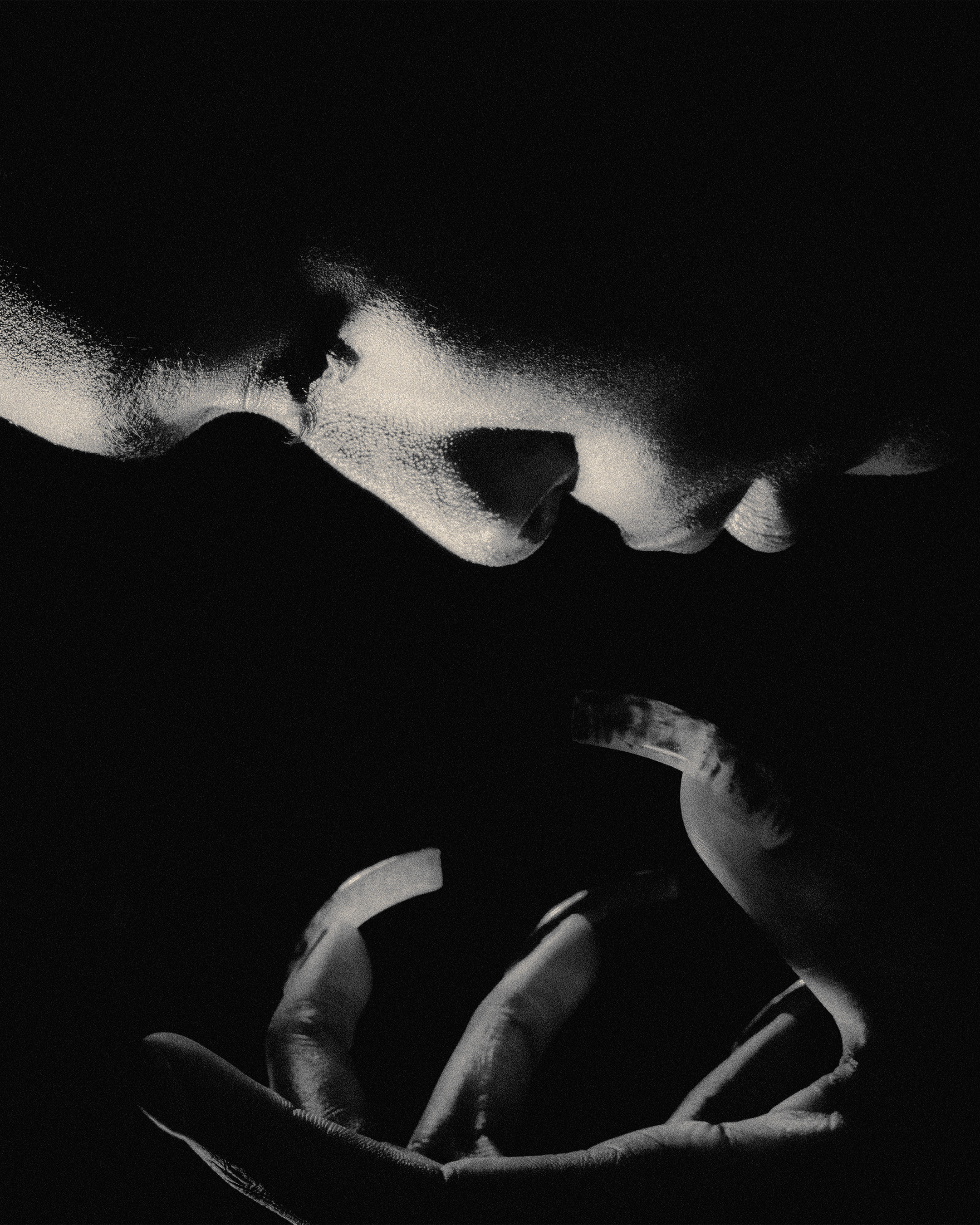 Monochromatic close-up of an adult's face and hands cupped together in darkness, lit dramatically to highlight skin texture.