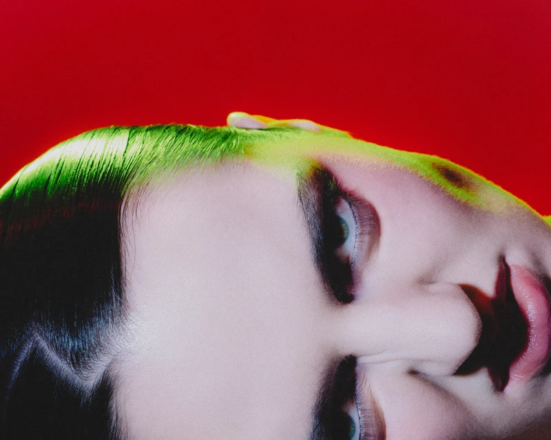 Close-up portrait of a woman lying down with vibrant green hair and red backdrop, focusing on her face and striking beauty.