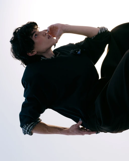 Young adult with dark curly hair and black shirt against white backdrop, arm raised, looking up, contemplative pose. Fashion portrait.