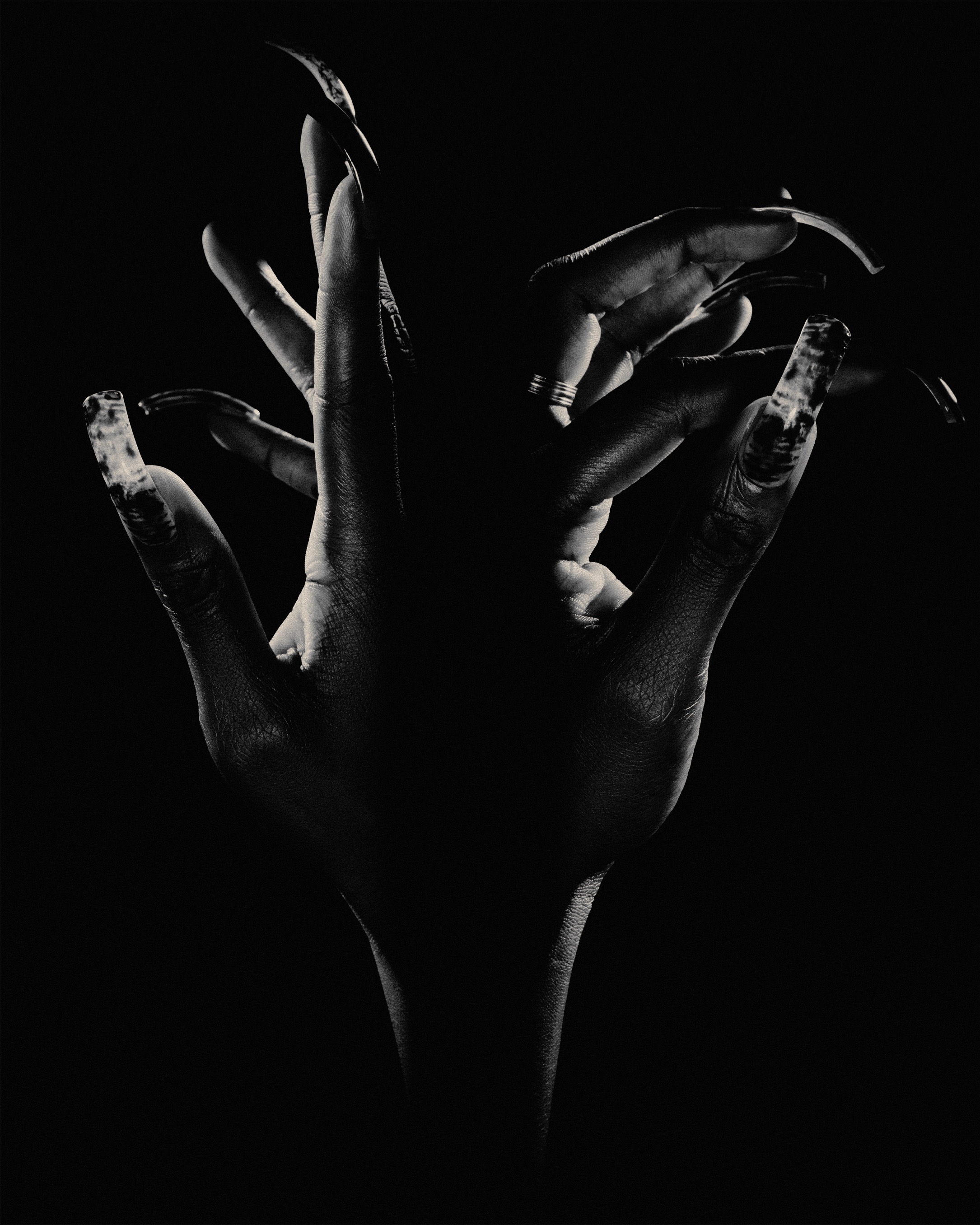 Close-up of two human hands with dark skin, long painted fingernails, and theatrical lighting against a solid black backdrop.