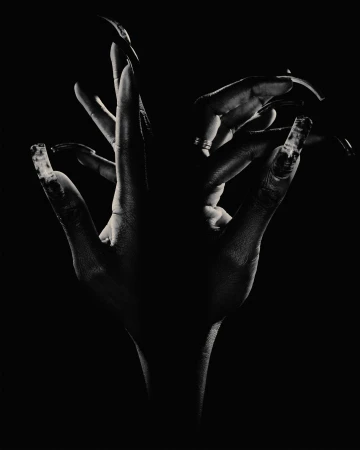 Close-up of two human hands with dark skin, long painted fingernails, and theatrical lighting against a solid black backdrop.