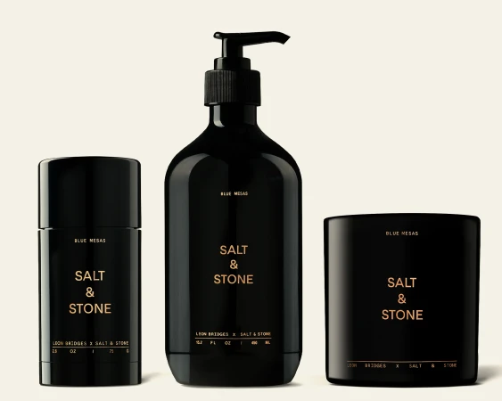 Three black Salt & Stone product containers on a light background: a deodorant, a pump bottle, and a candle. Minimalist product photography.