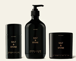 Three glossy black cosmetic containers from Salt and Stone x Leon Bridges including a pump bottle, candle, and deodorant on a pale background.