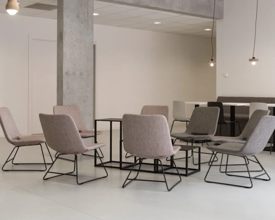 Modern office break room with neutral-toned chairs arranged around small tables, set against a backdrop of white walls and concrete pillars.