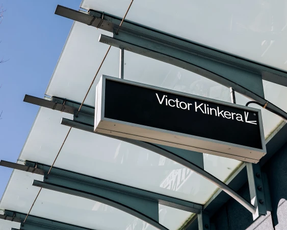 Low-angle view of a black sign reading 'Victor Klinkera' hanging from a metal structure with a translucent awning above on a sunny day.