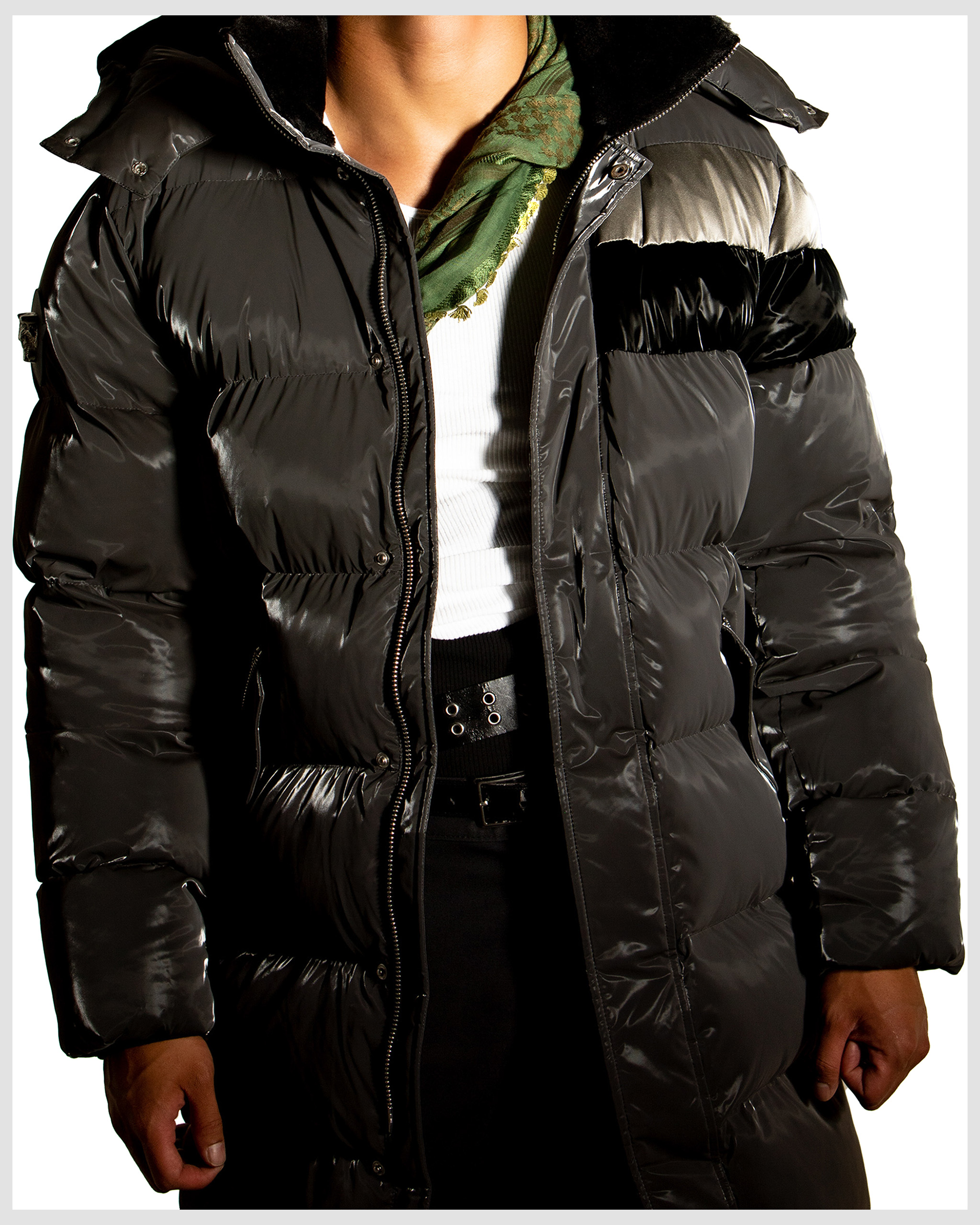 Person in long grey puffer jacket with black accents and a green scarf, posing in a bright studio setting. Fashion.