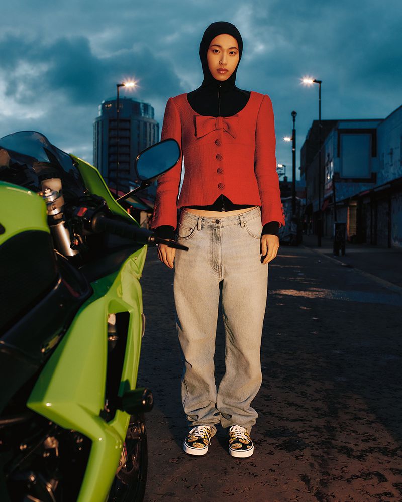 A person stands next to a Kawasaki motorcycle wearing a red blazer, blue jeans, and a black head covering on an empty street.
