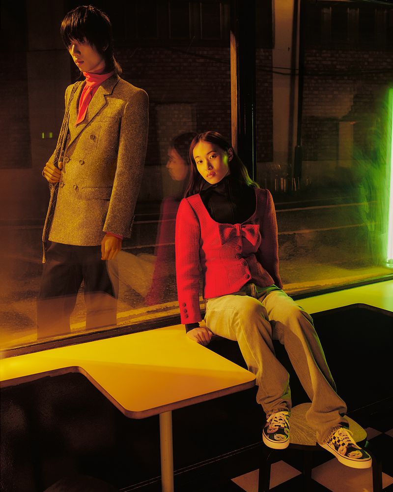 Two people in an indoor location with warm lighting. A woman sits on a table in a red jacket, while a man stands in the background.