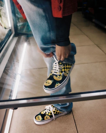 Person in light-wash jeans tying the white laces of black, yellow, and white patterned Vans sneakers with yellow cartoon faces.