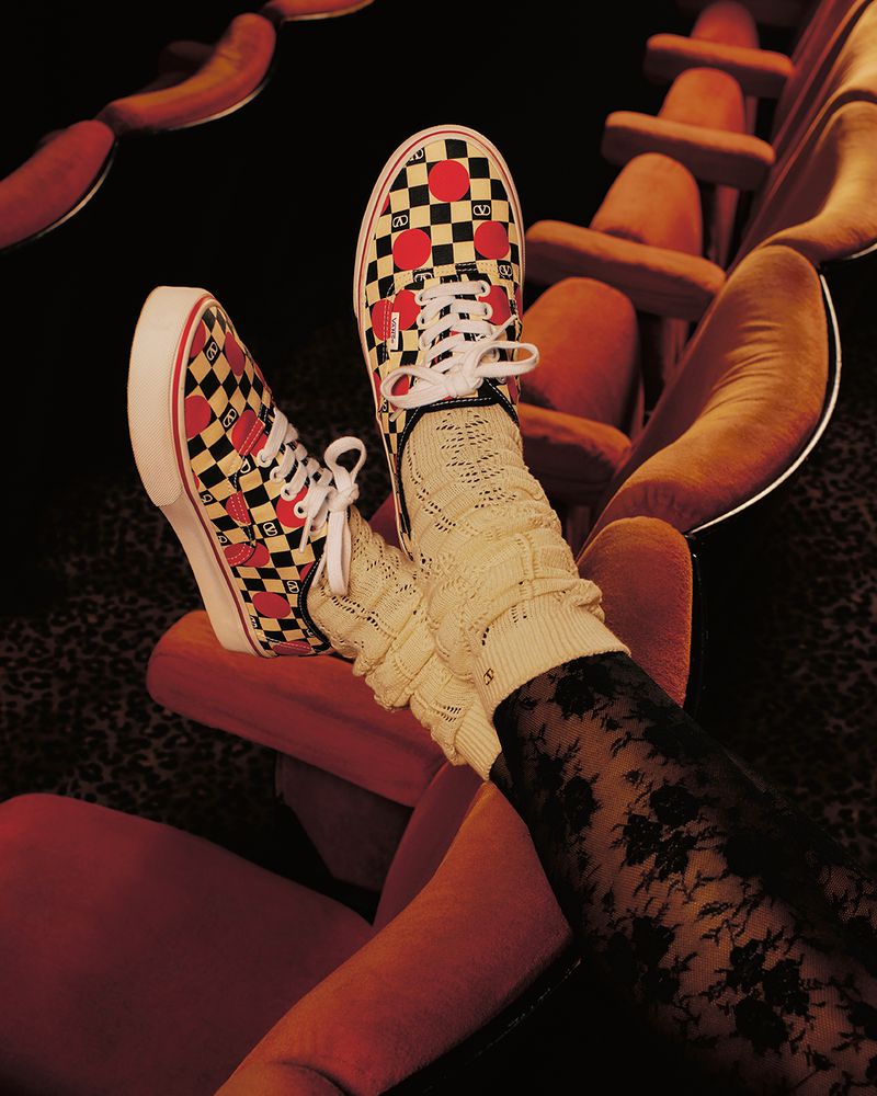 A pair of checkerboard Vans sneakers with red hearts on a red movie theater seat, with a person wearing patterned socks.