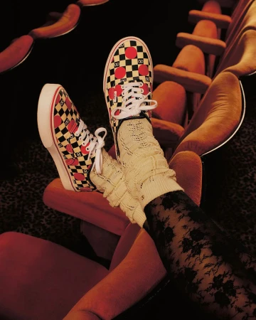A pair of checkerboard Vans sneakers with red hearts on a red movie theater seat, with a person wearing patterned socks.