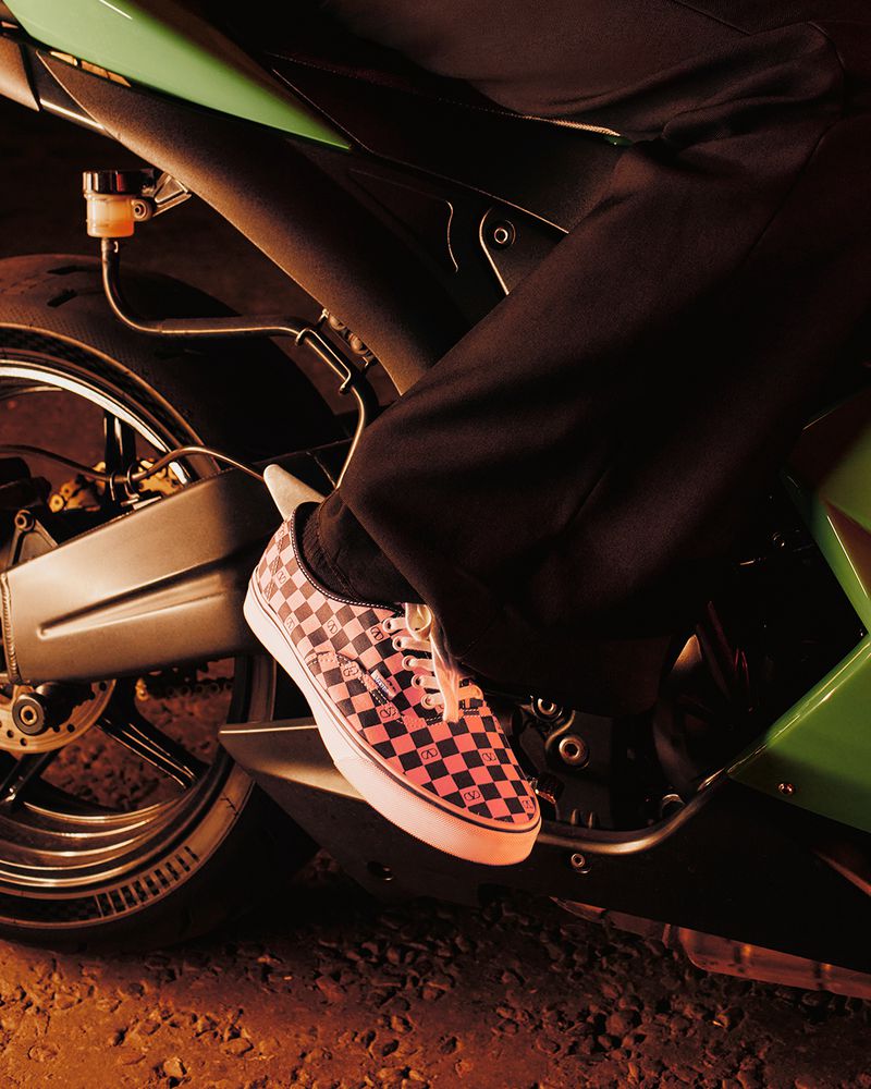 Low angle shot of a person on a motorcycle with pink and black checkerboard sneakers, dark pants, and a green motorcycle.
