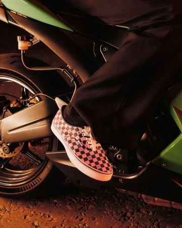 Low angle shot of a person on a motorcycle with pink and black checkerboard sneakers, dark pants, and a green motorcycle.