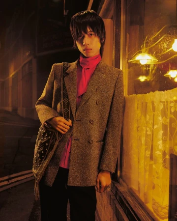 Portrait of a young adult with a gray blazer and bag standing by a yellow window at night in an urban outdoor setting.