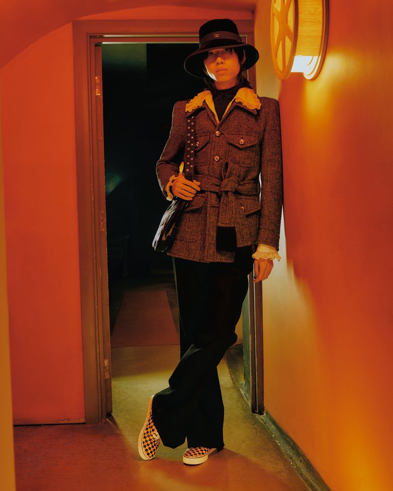 Portrait of a stylish woman in a tweed jacket, black trousers, and checkered shoes, leaning against an orange wall indoors with warm lighting.