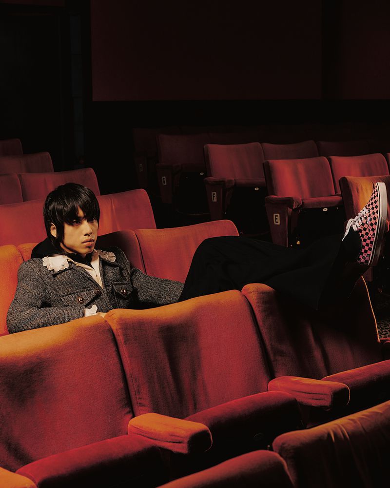 Young adult relaxes in red theater seats wearing a grey tweed jacket and checkered shoes, backlit by dramatic lighting from the screen.