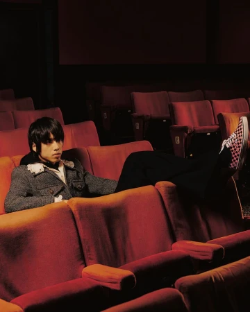 Young adult relaxes in red theater seats wearing a grey tweed jacket and checkered shoes, backlit by dramatic lighting from the screen.