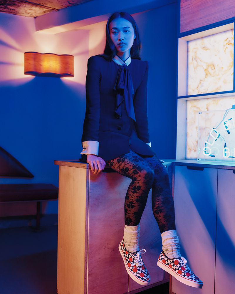 A slender woman in a suit jacket sits on a cabinet wearing patterned leggings and sneakers in a blue-lit indoor setting with a lamp.